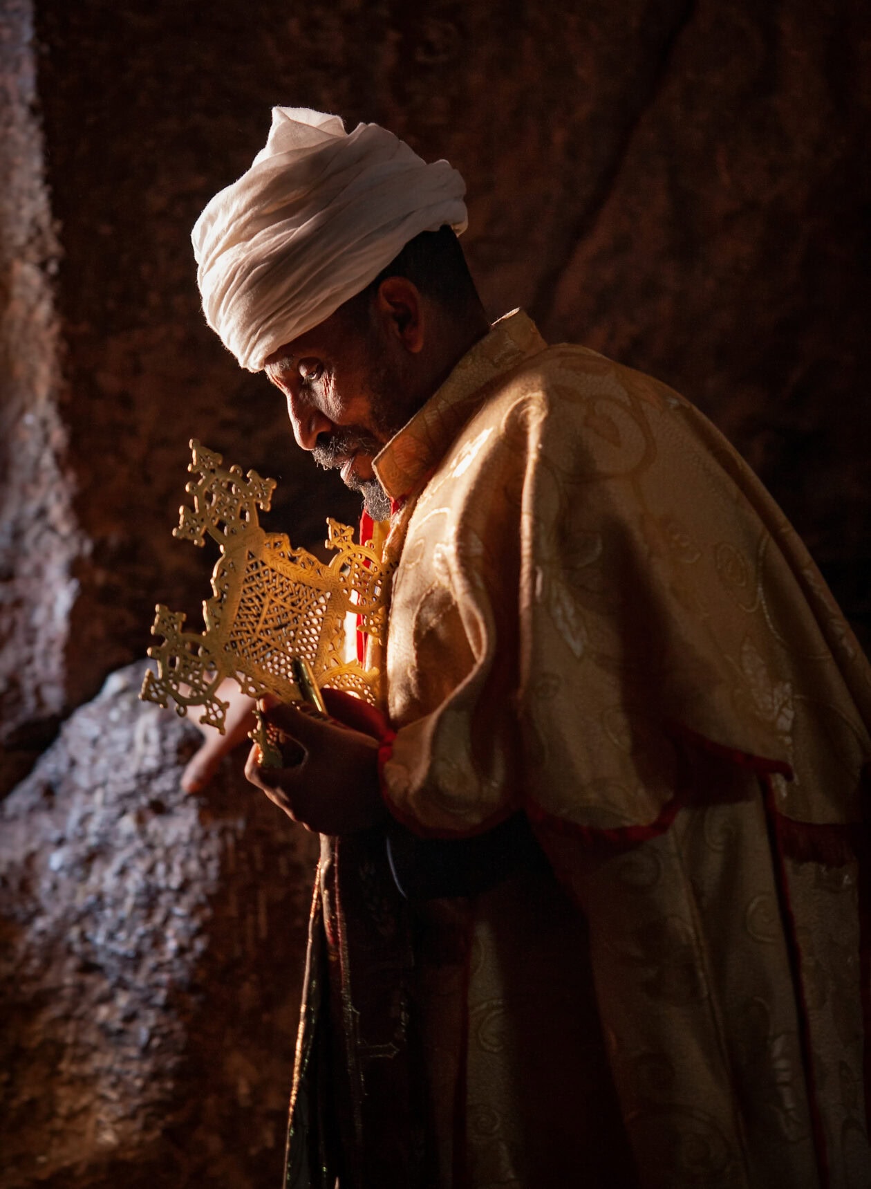 Etiopský ortodoxní kněz s procesním křížem ve skalním otvoru v ranním světle, Lalibela, Etiopie